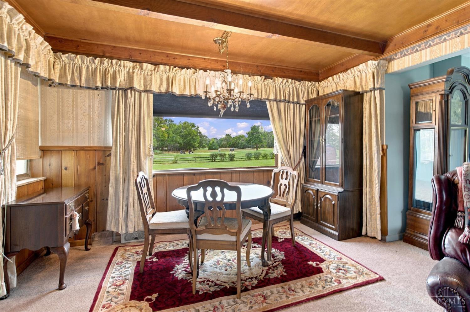 Dining room with wood-paneled ceiling, round table, chandelier, and a wide window overlooking a green lawn.