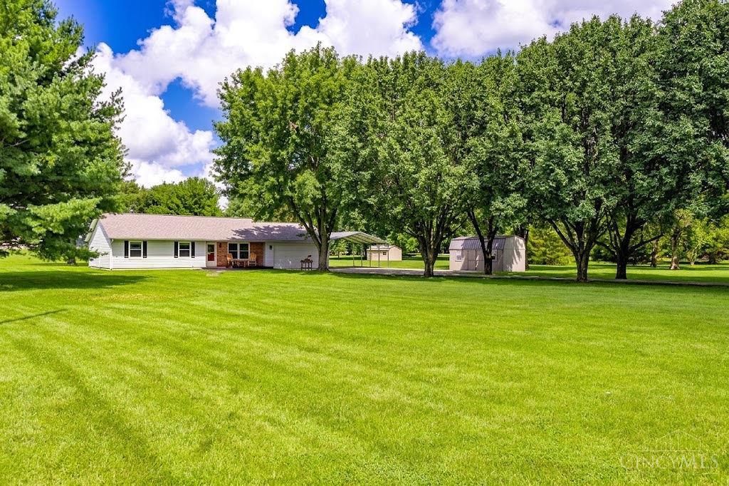 Single-story home beyond a broad lawn, with mature trees, a carport, and a detached shed.