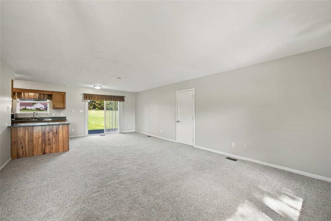 Vacant living room with gray carpet, wood kitchen cabinets, and a sliding glass door to the lawn.