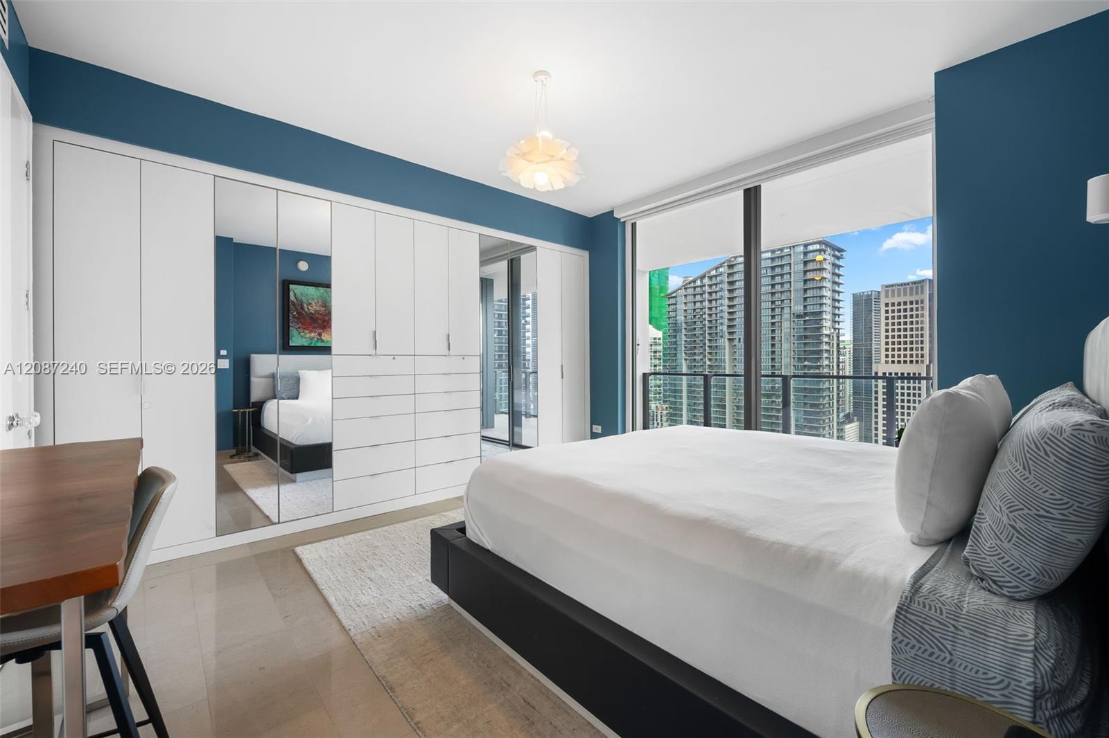 Modern bedroom with mirrored wardrobes, blue walls, and a city view through floor-to-ceiling windows.