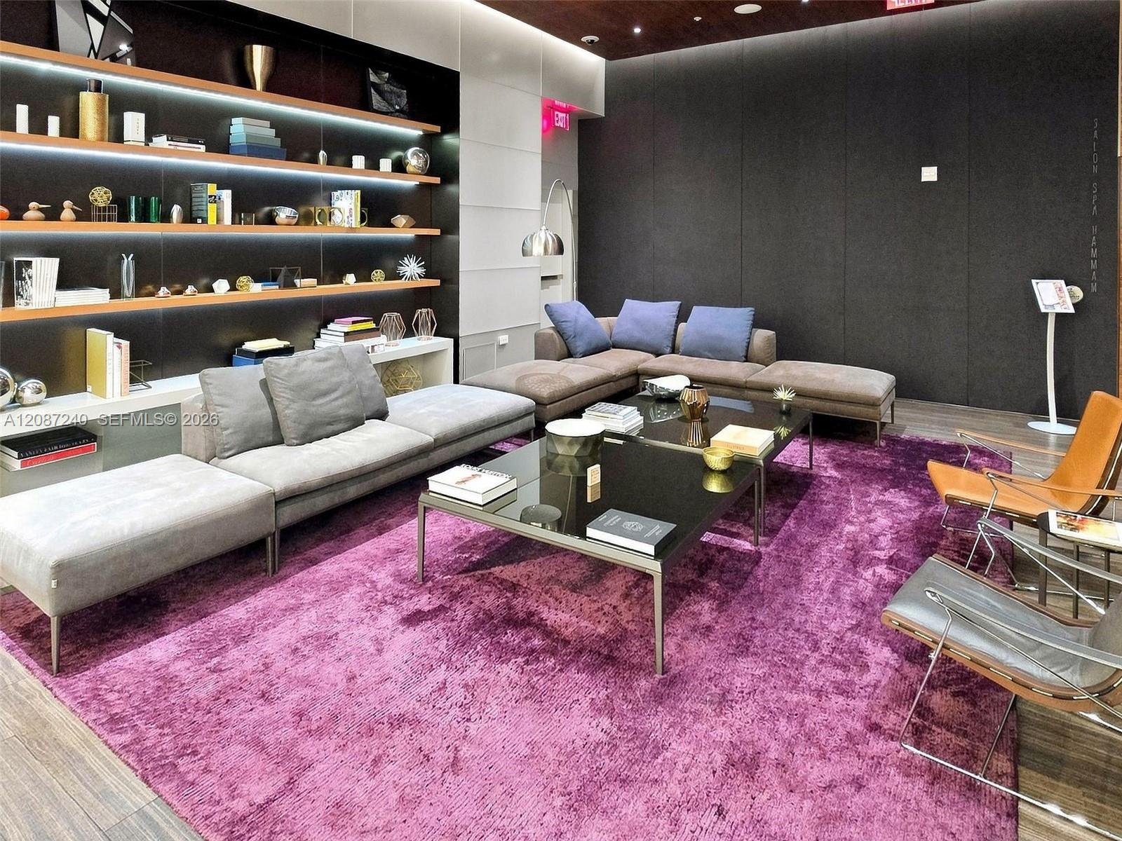 Furnished lounge with gray sectional sofas, illuminated display shelves, and a vivid purple rug.