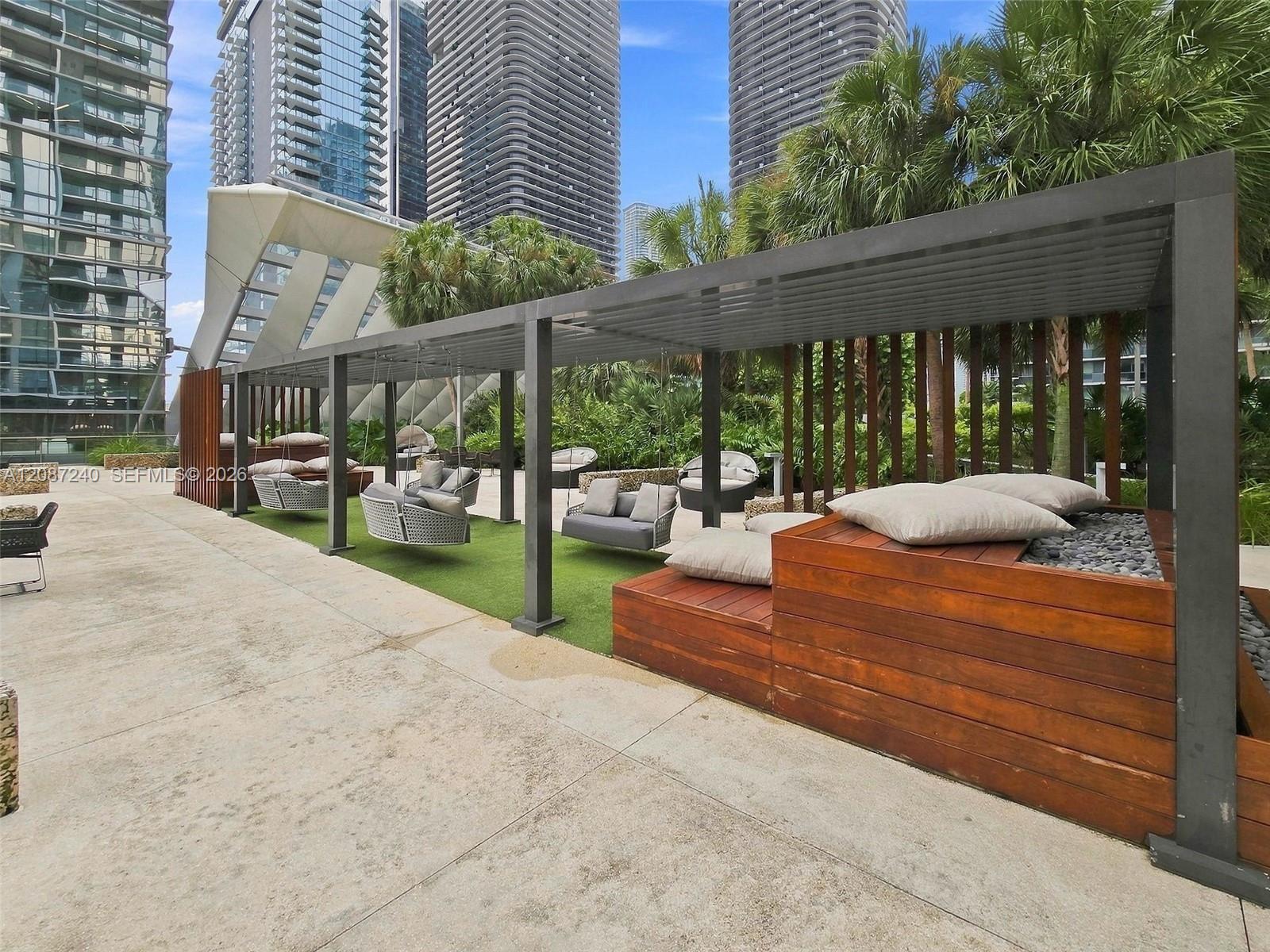 Condominium terrace with a pergola, cushioned lounge seating, tropical landscaping, and high-rise buildings.