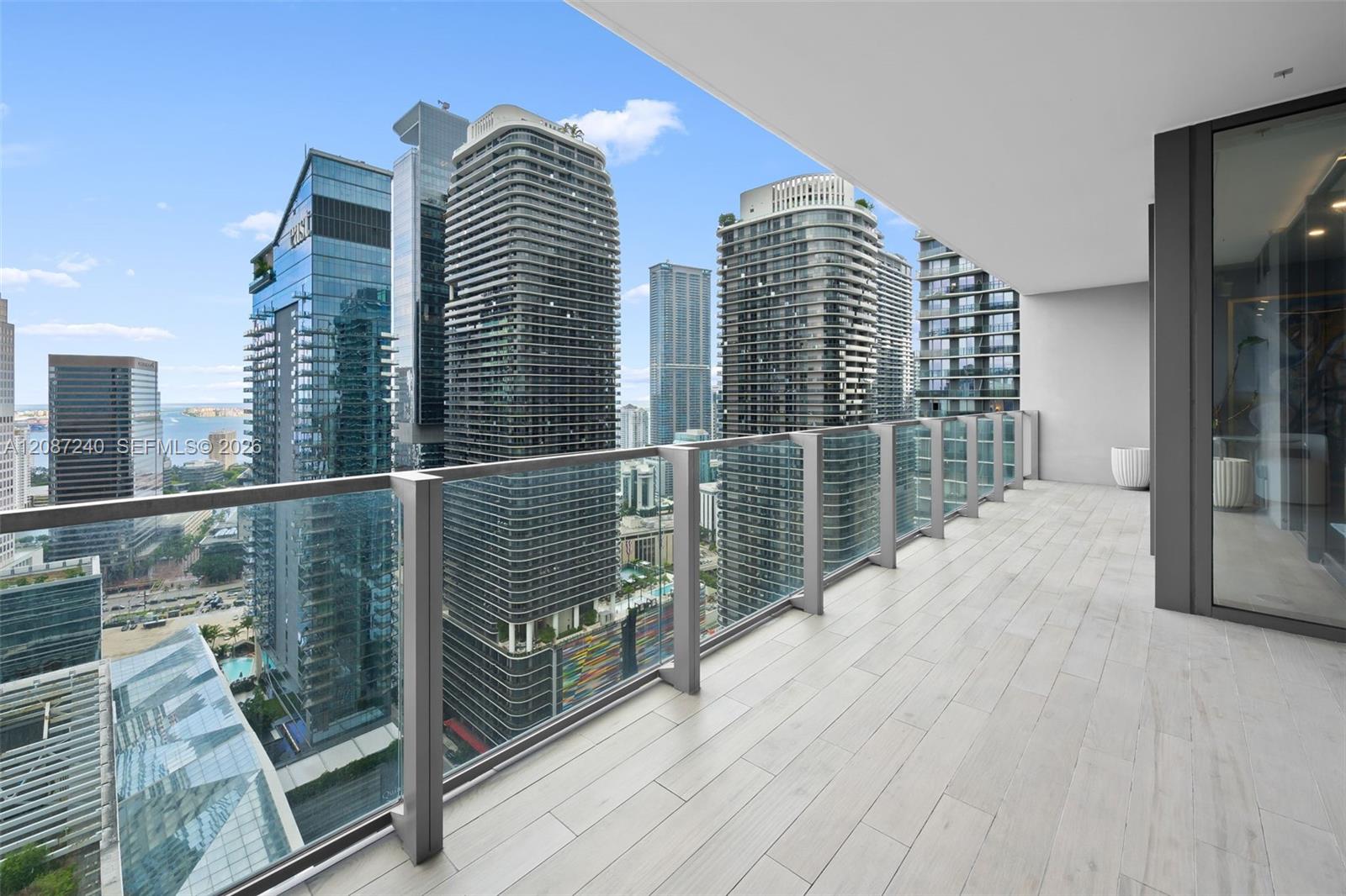 High-rise condo balcony with glass railing overlooking downtown Miami towers and distant water.