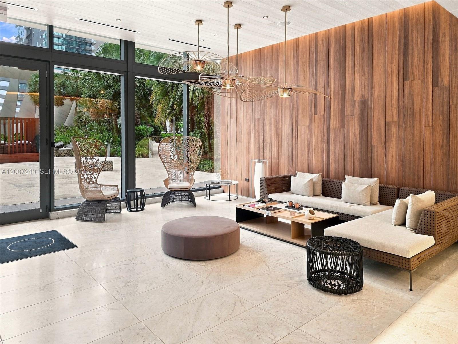 Condominium lobby lounge with woven seating, a wood-paneled wall, and windows overlooking tropical landscaping.