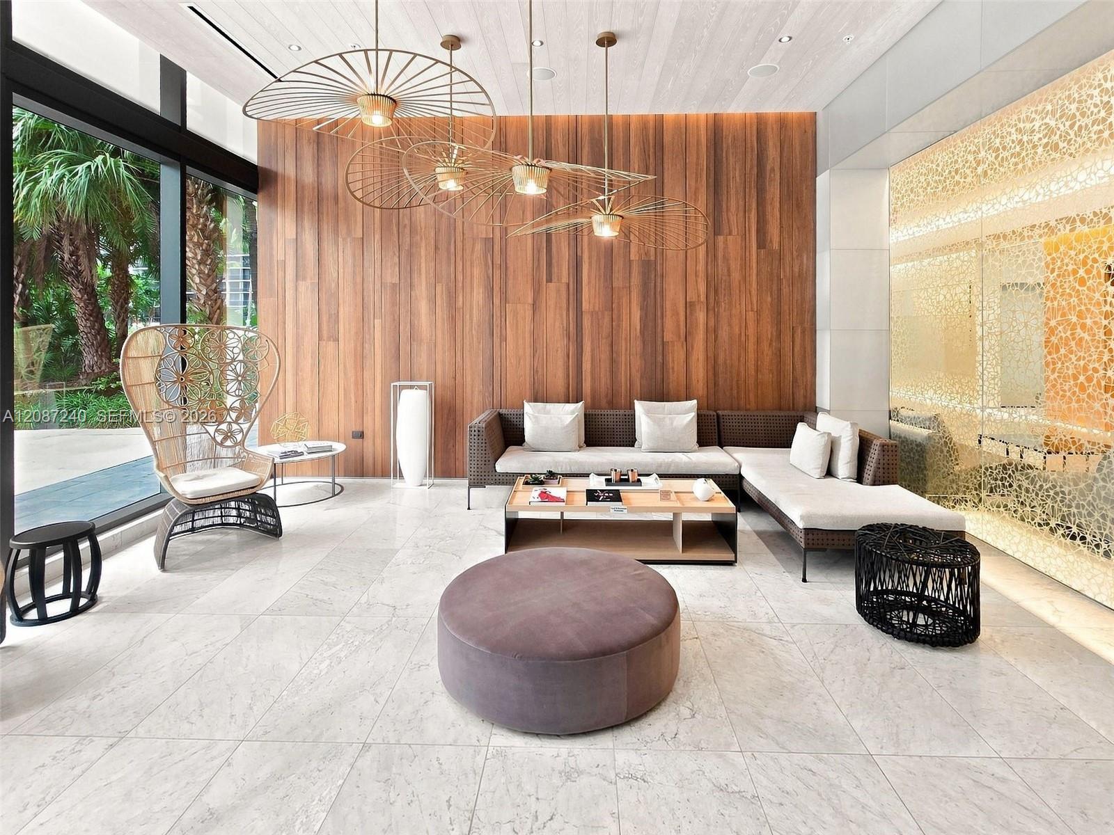 Condominium lobby lounge with a wood-paneled wall, sculptural pendant lights, and windows overlooking palm trees.