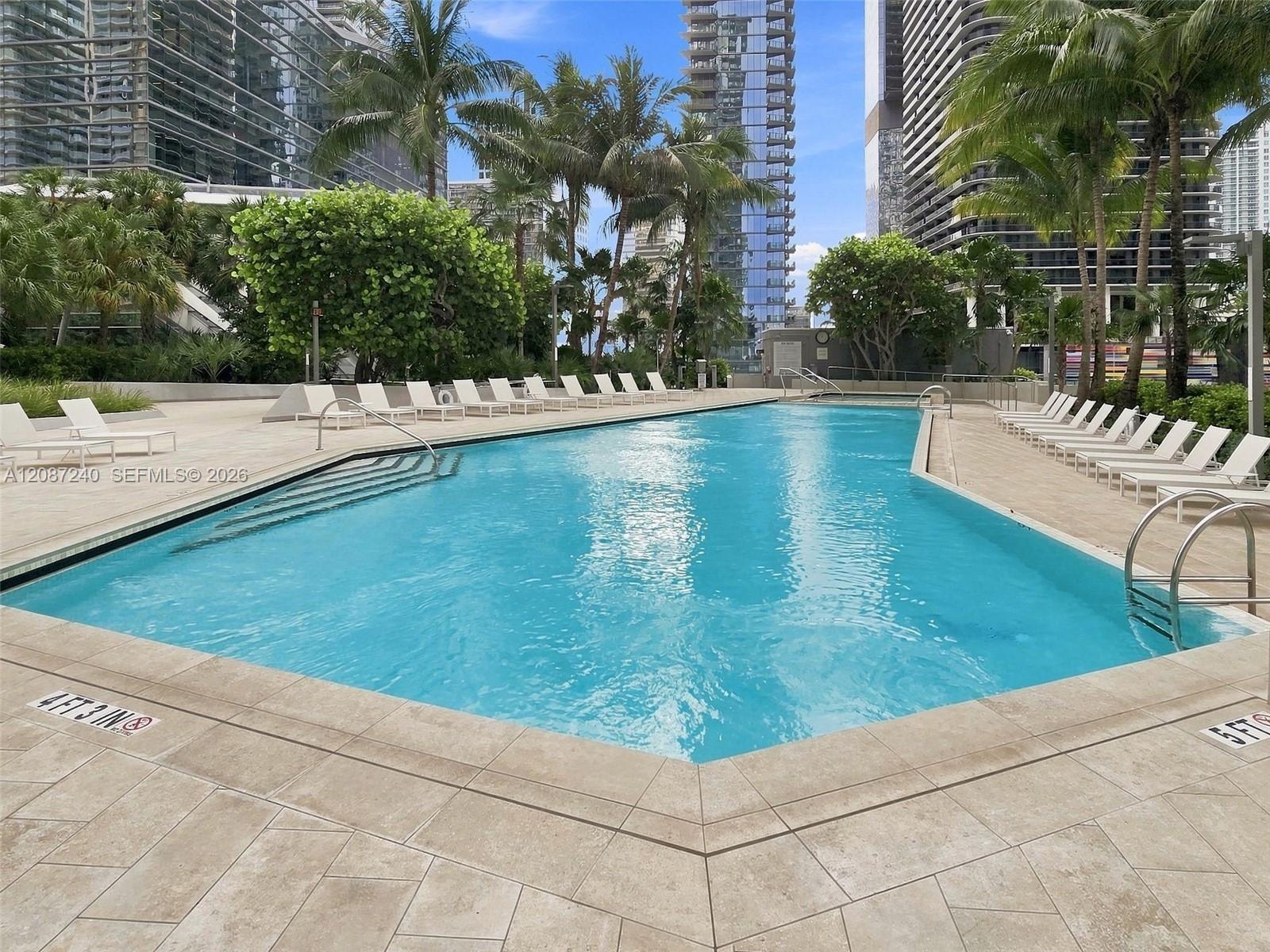 Outdoor condominium pool with turquoise water, tiled deck, lounge chairs, and palm trees among high-rises.