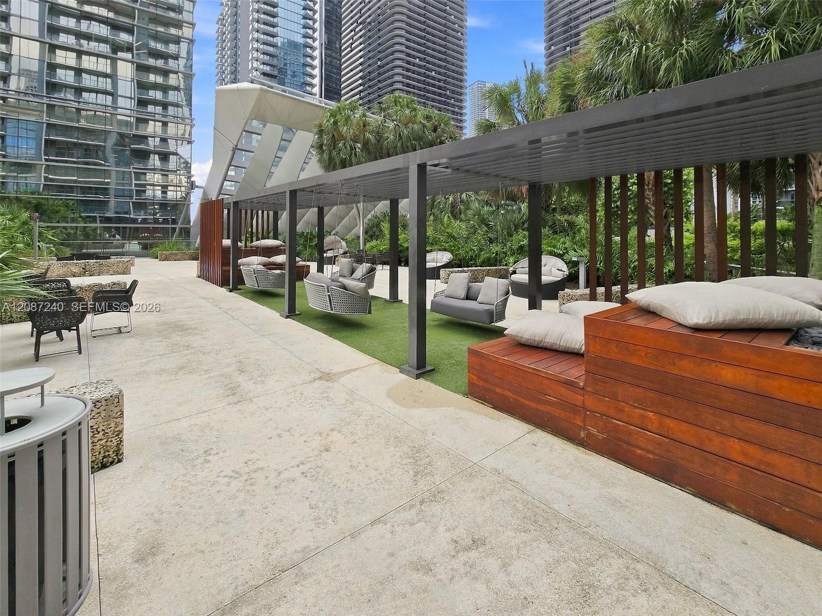 Condominium terrace with cushioned lounge seating beneath a pergola, surrounded by palms and high-rise buildings.