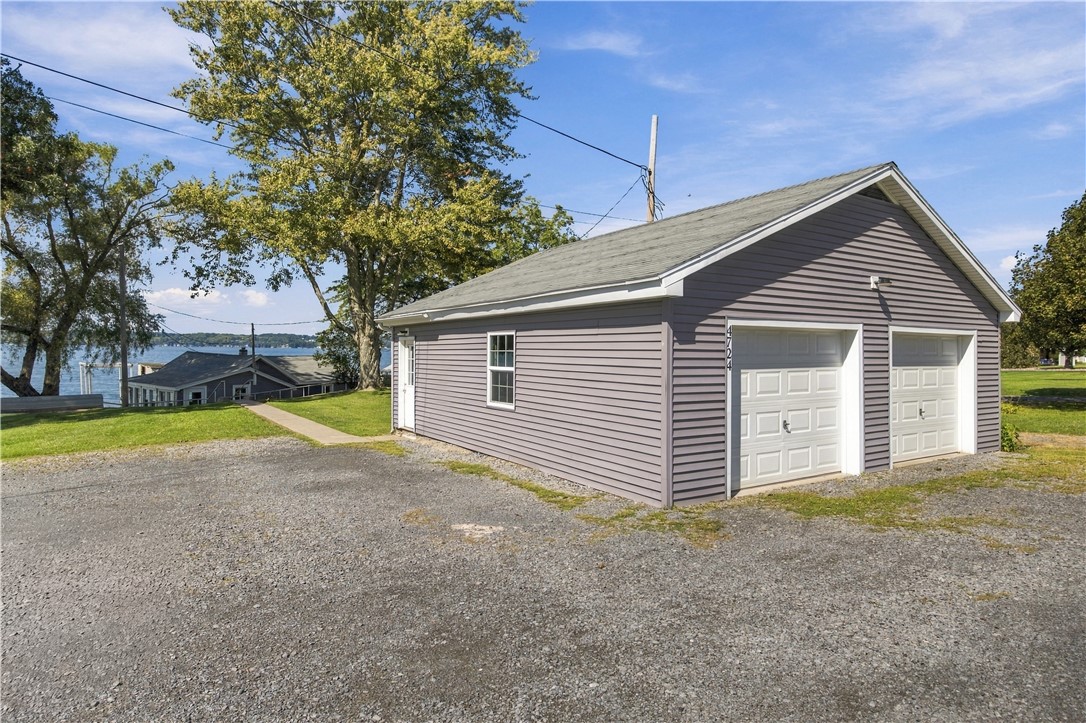 Detached gray two-door garage beside a gravel driveway, with a lake visible behind it.