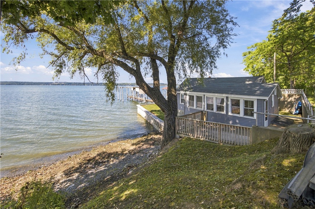 Gray lakeside cottage beside a dock, shaded by mature trees along a rocky shoreline.