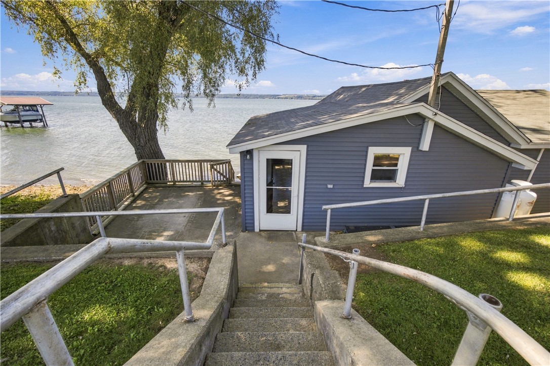 Gray lakeside home beside a shaded concrete patio, with a covered dock visible across the water.