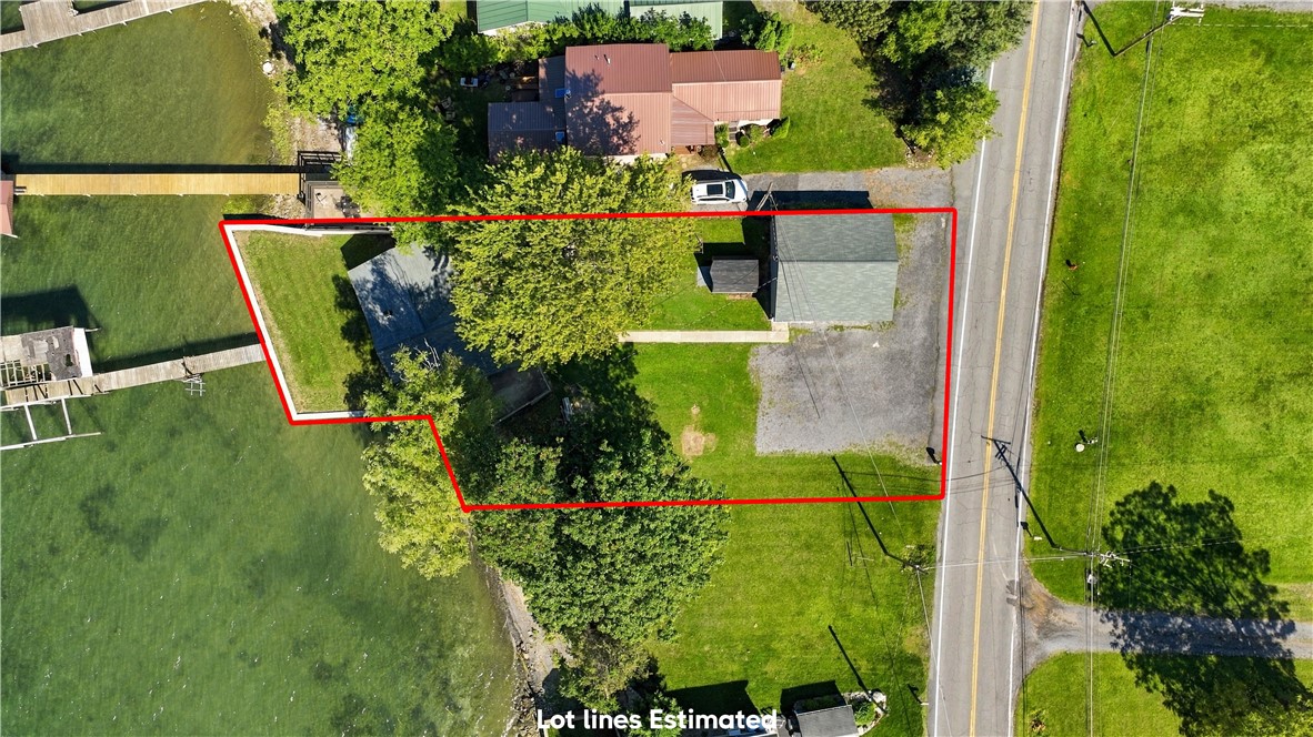 Aerial view of a waterfront lot outlined in red, with a house, outbuilding, lawn, and gravel parking area.