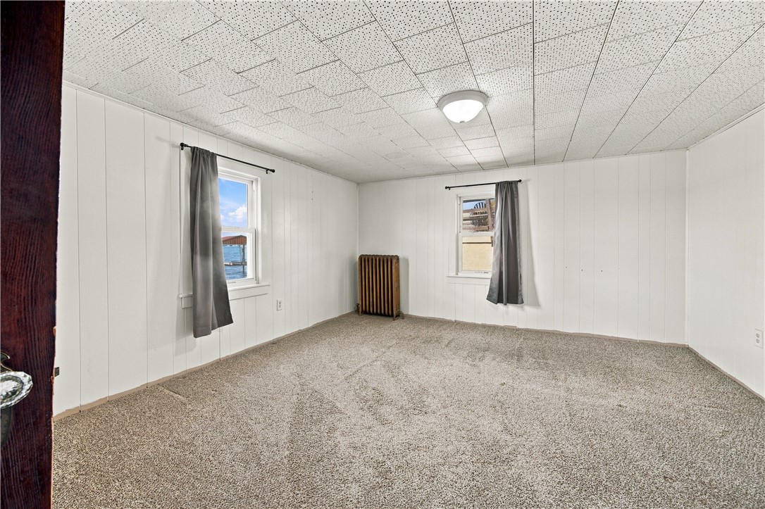 Empty carpeted room with white paneled walls, curtained windows, and a radiator.