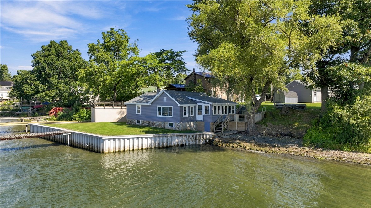 Gray lakefront house with a stone foundation, wooden shoreline retaining wall, and mature trees.