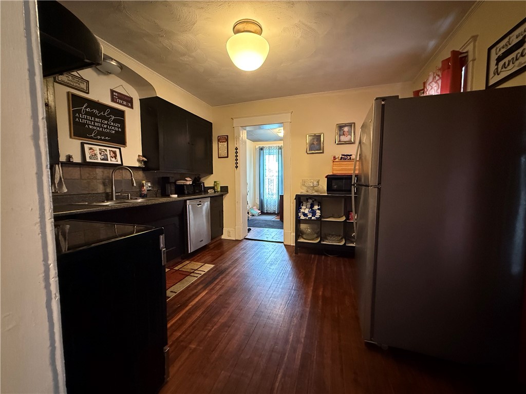 Kitchen with dark cabinets, a stainless-steel dishwasher, wood floors, and a doorway to another room.
