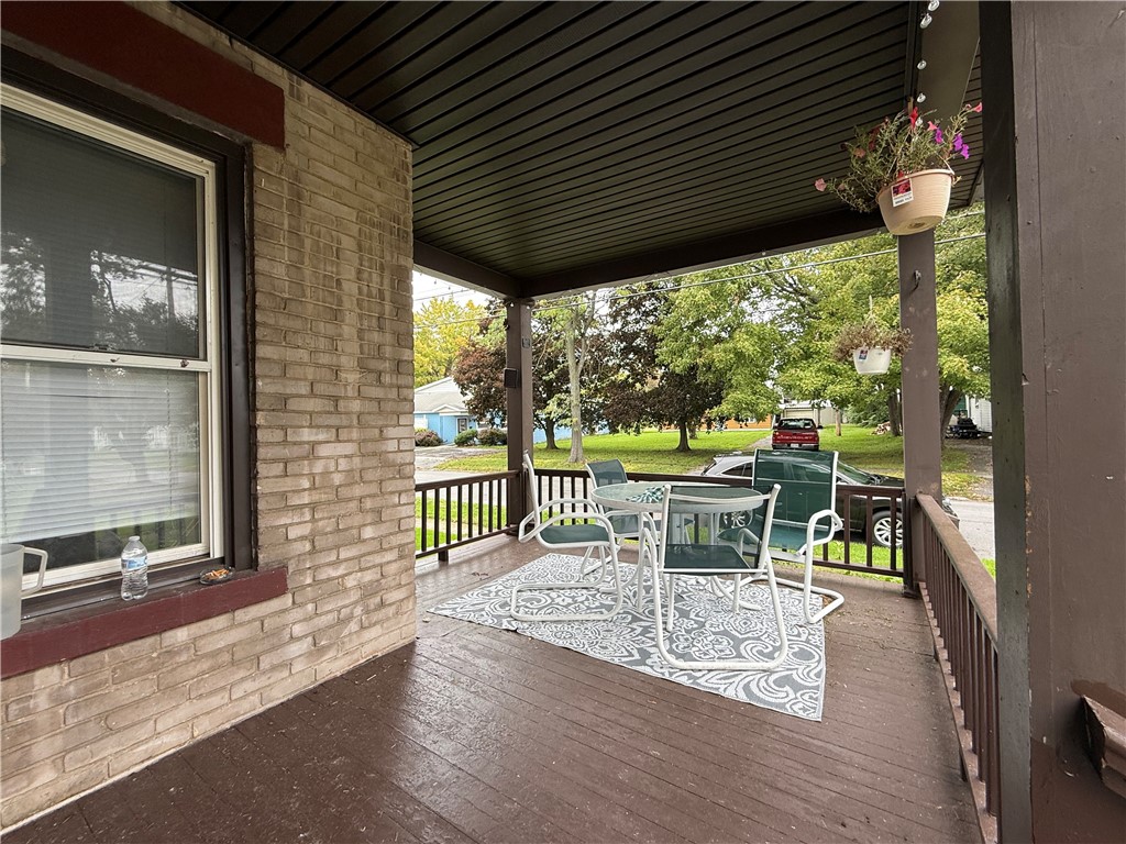 Covered front porch with white patio furniture, hanging flowers, and a leafy street view.