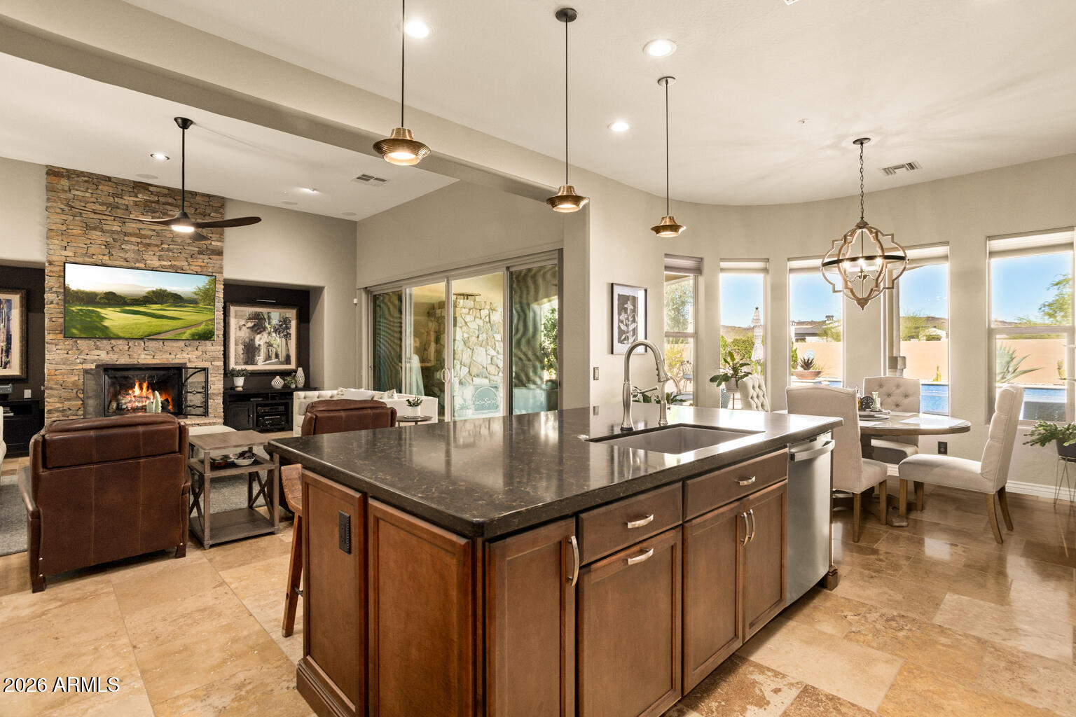 Open kitchen with a large stone island, stone fireplace, and tall windows overlooking a pool.