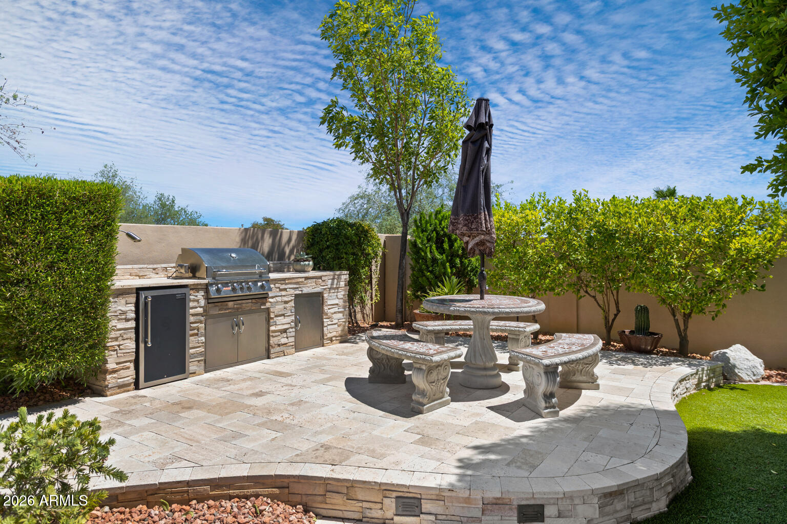 Backyard patio with a stone outdoor kitchen, stainless steel grill, and round stone table with benches.