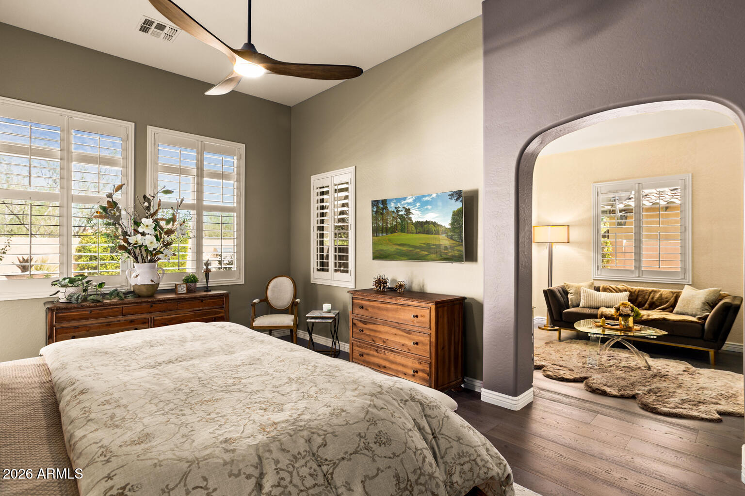 Furnished bedroom with plantation shutters, wooden dressers, and an arched opening to a sitting room.