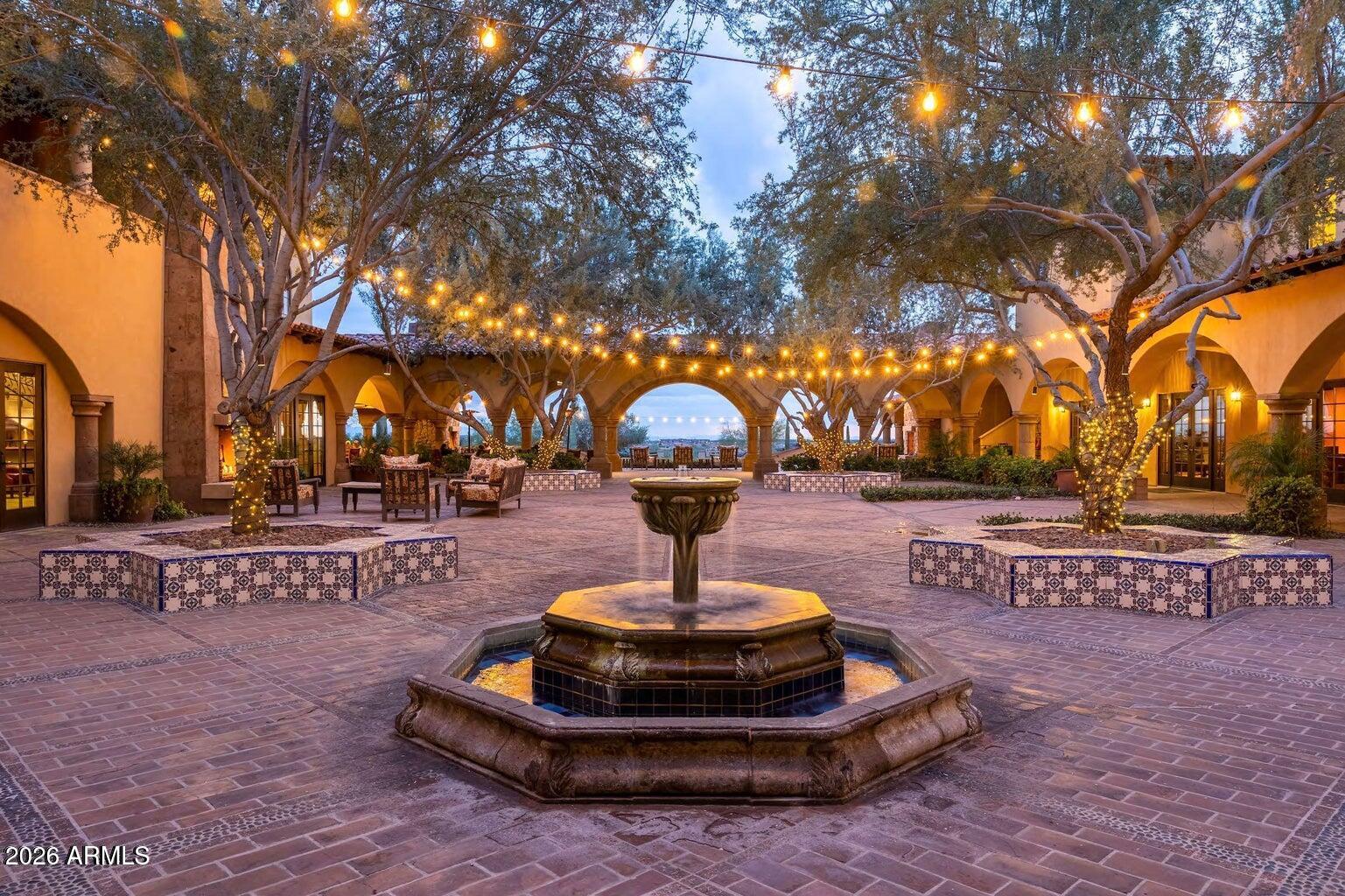 Courtyard with a stone fountain, arched walkways, mature trees, and glowing string lights.
