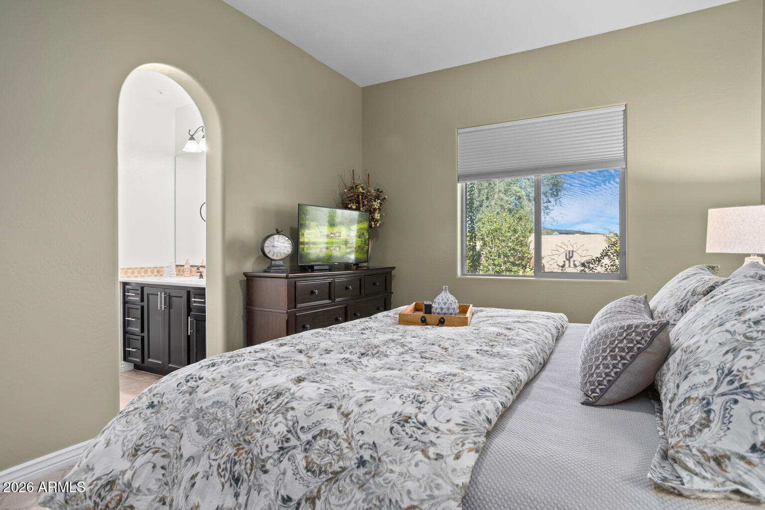 Furnished bedroom with a patterned bed, dark wood dresser, television, and arched vanity opening.