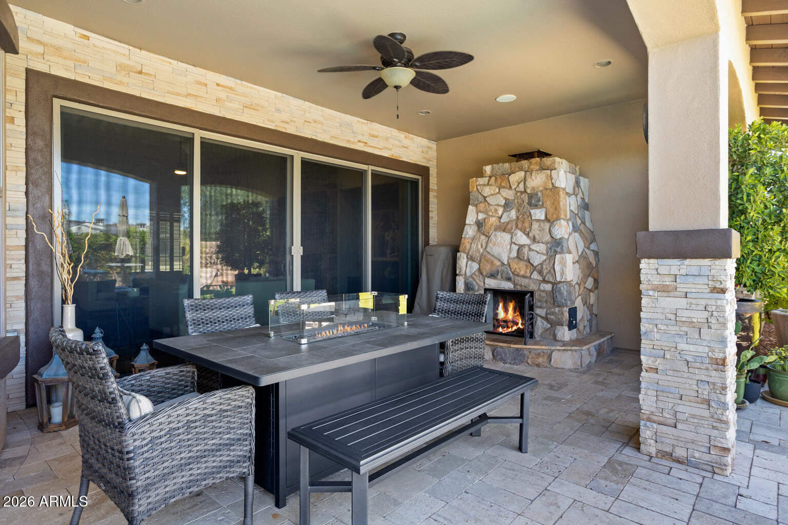 Covered stone patio with outdoor dining furniture and a lit stone fireplace.