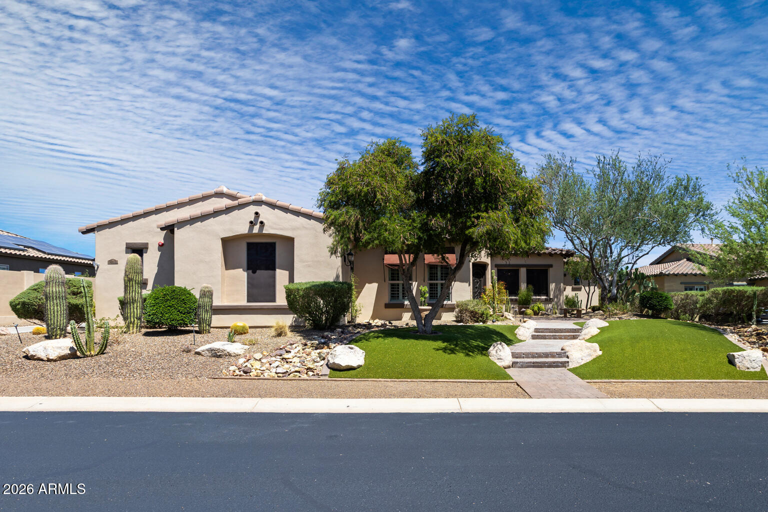 Single-story stucco home with desert landscaping, mature trees, and a green lawn along the front walkway.