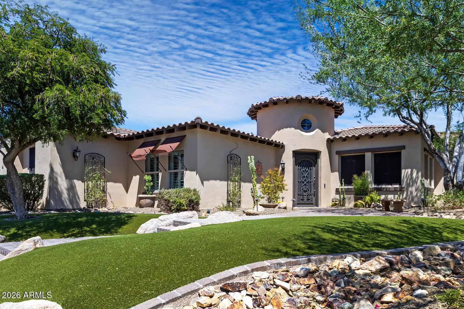 Stucco home with a round tower, decorative entry door, green lawn, and mature trees.