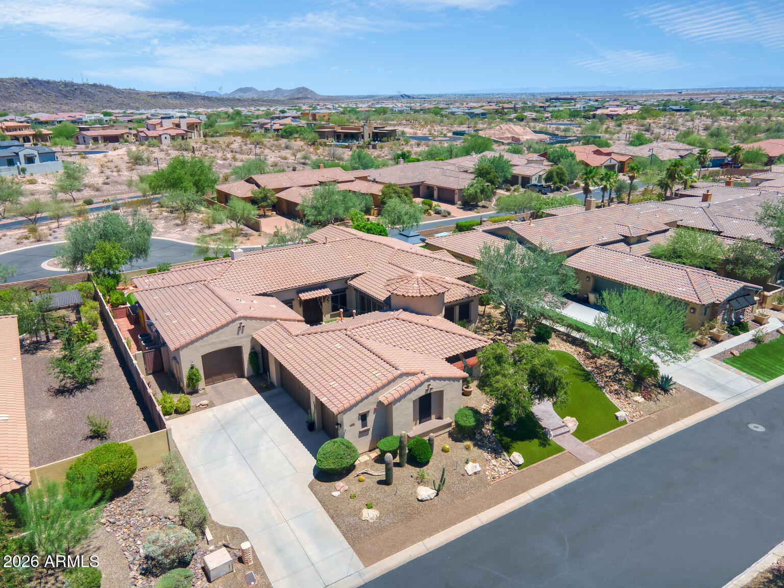 Aerial view of a tile-roofed home with desert landscaping, neighboring homes, and distant hills.