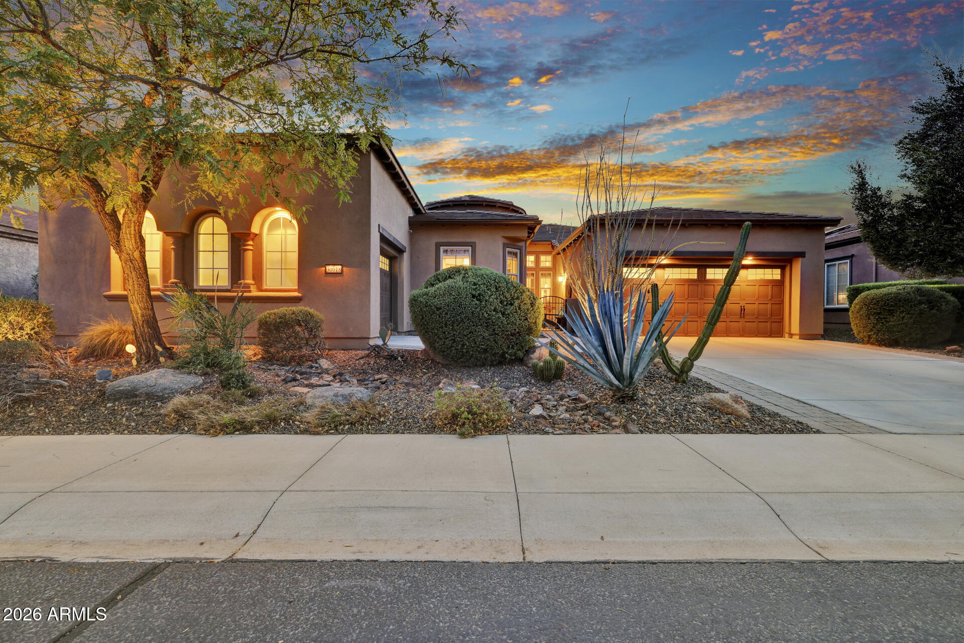 Stucco home with arched windows, an illuminated garage, and desert landscaping at dusk.