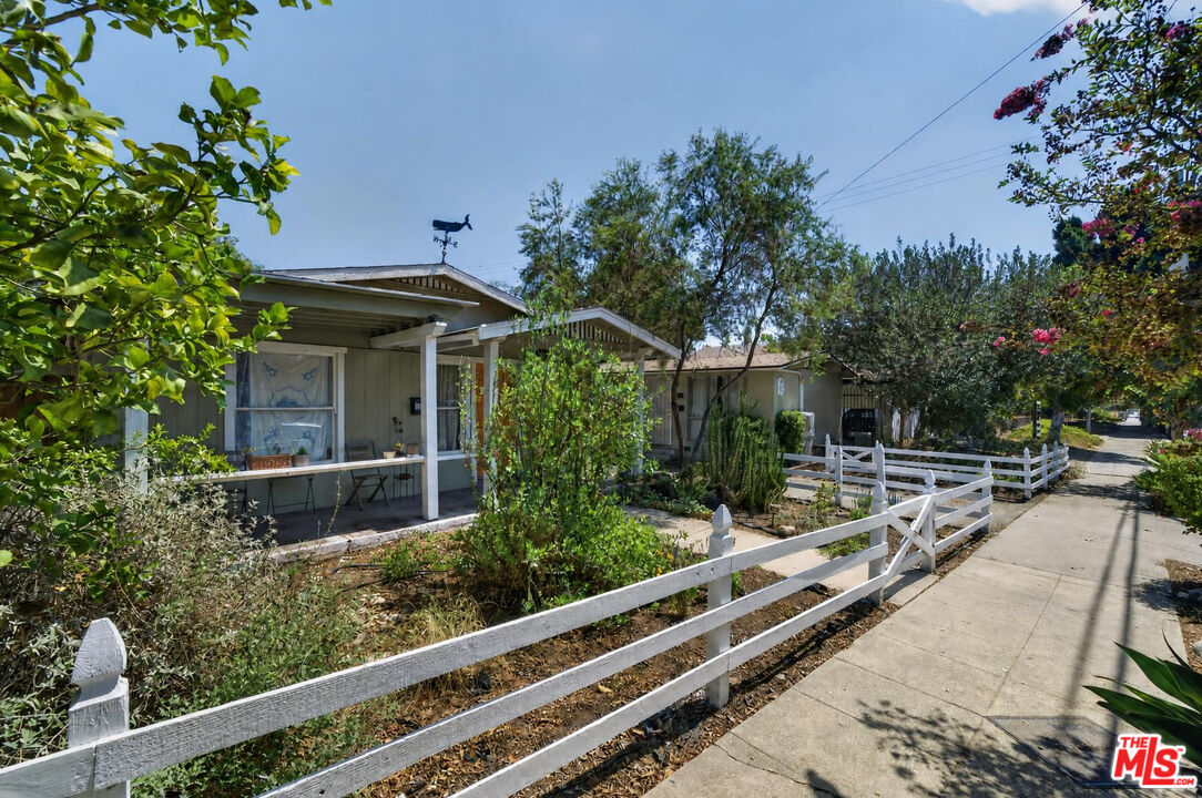 Single-story home with a covered porch, leafy garden, and white fence along the sidewalk.
