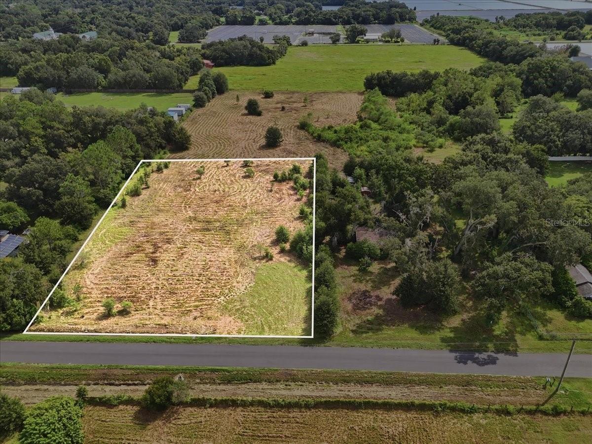 Aerial view of a cleared vacant lot outlined in white beside a road, surrounded by trees and fields.