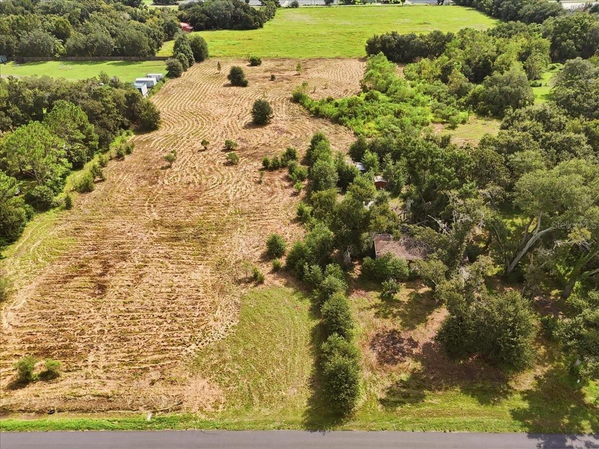 Aerial view of a cleared parcel bordered by trees, green fields and a paved road.