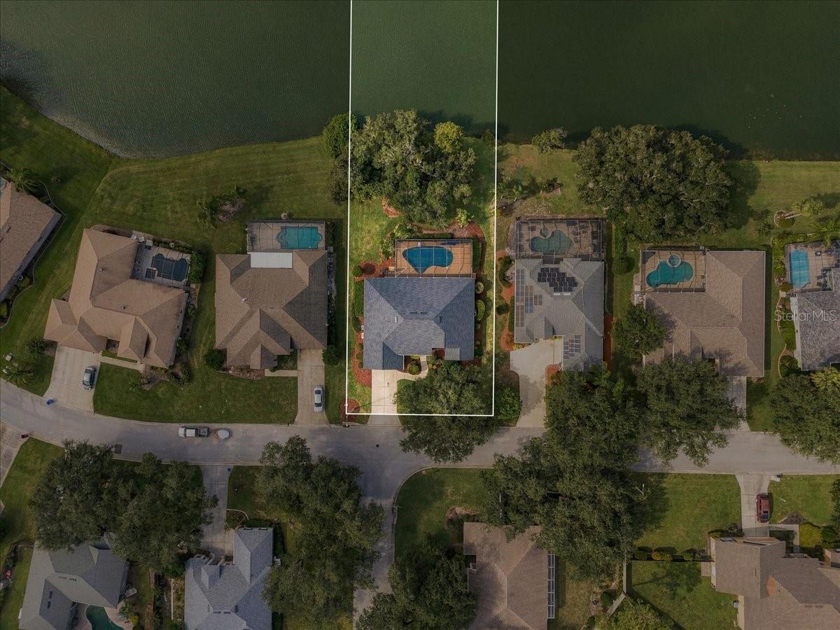 Aerial view of a lakefront home with a backyard pool and outlined lot, surrounded by neighboring houses.