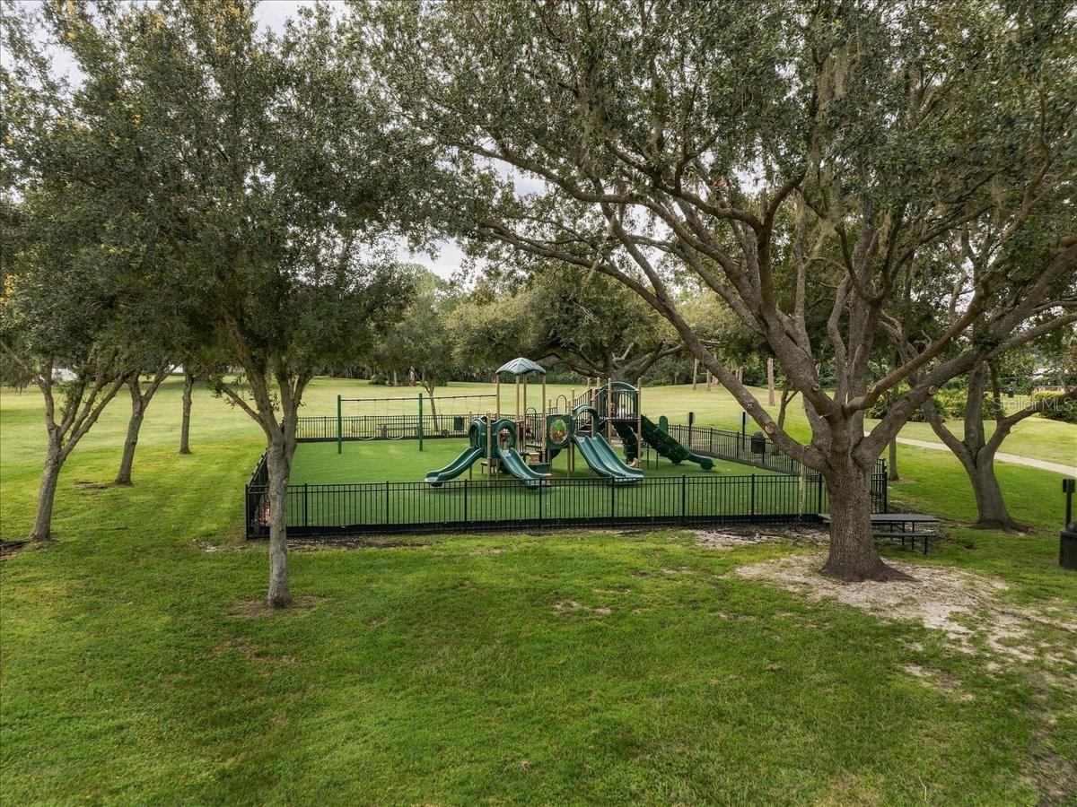 Fenced playground with green slides beneath mature shade trees.
