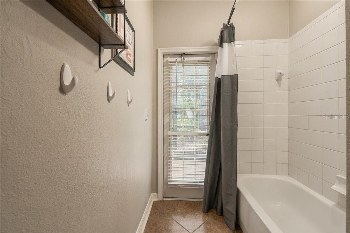 Bathroom with a white bathtub, tiled surround, and a gray shower curtain beside a glass door with blinds.