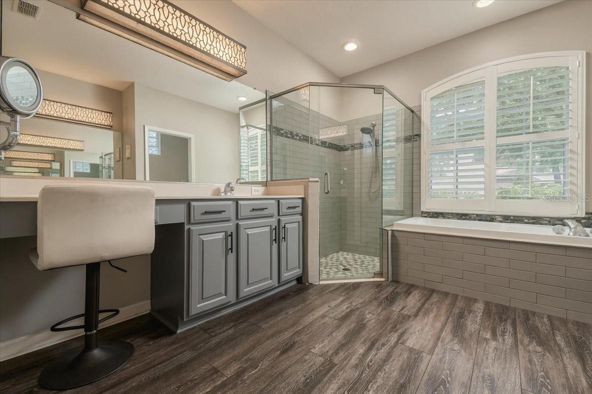 Primary bathroom with gray vanity, glass shower, and bathtub beneath plantation-shuttered windows.