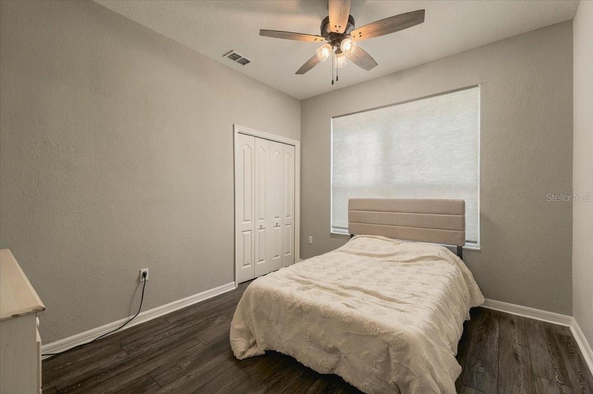 Bedroom with a bed beneath a wide window, ceiling fan, double closet doors, and dark wood flooring.