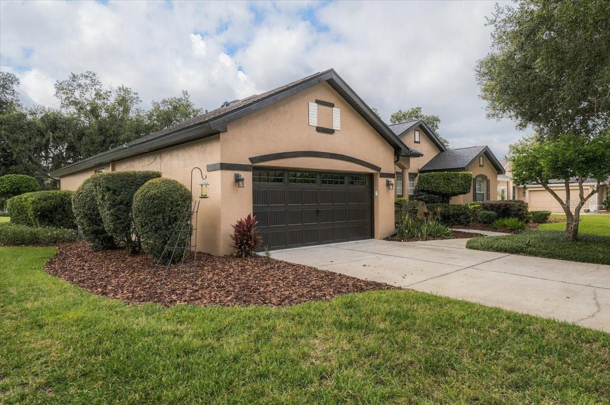 Beige stucco home with a dark double garage door, concrete driveway, and landscaped front lawn.