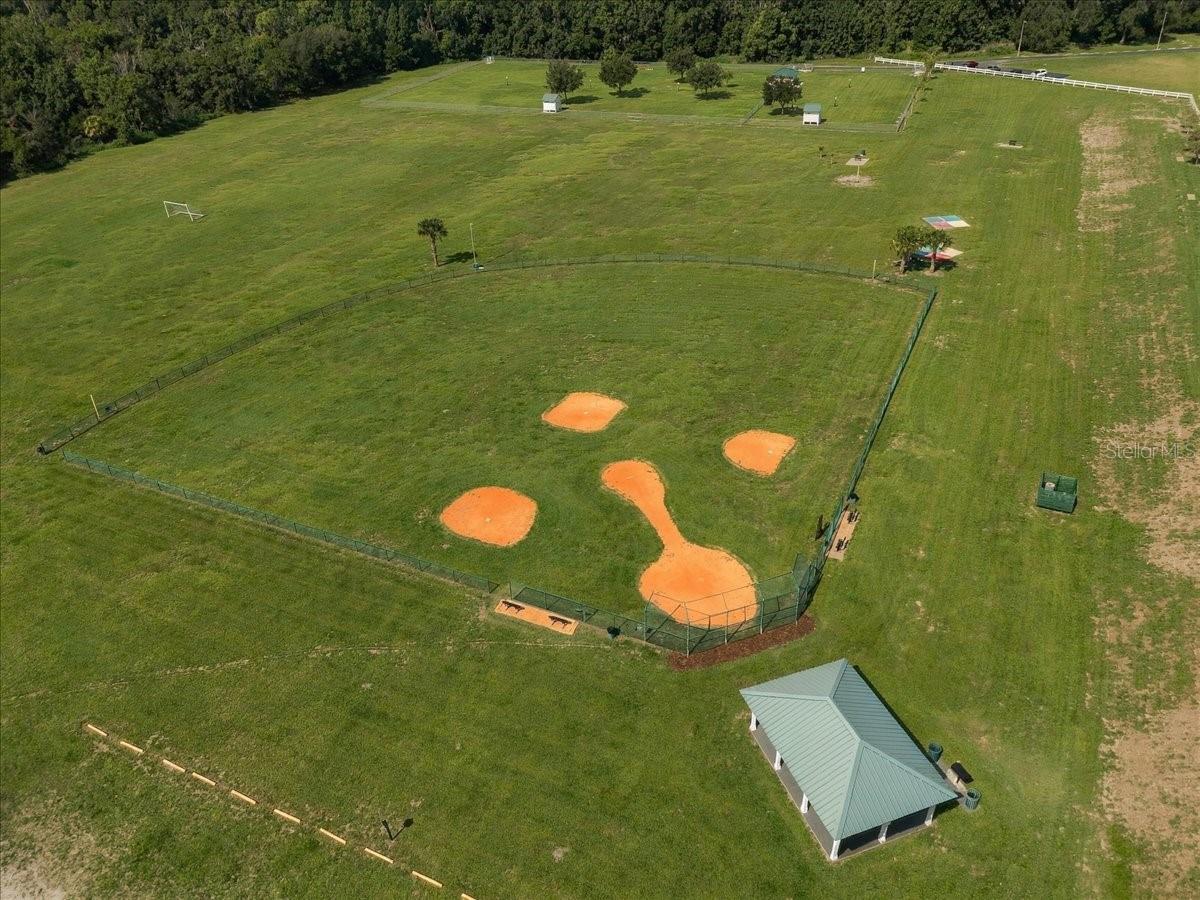 Fenced baseball field with orange dirt bases set within broad grassy grounds and a nearby pavilion.