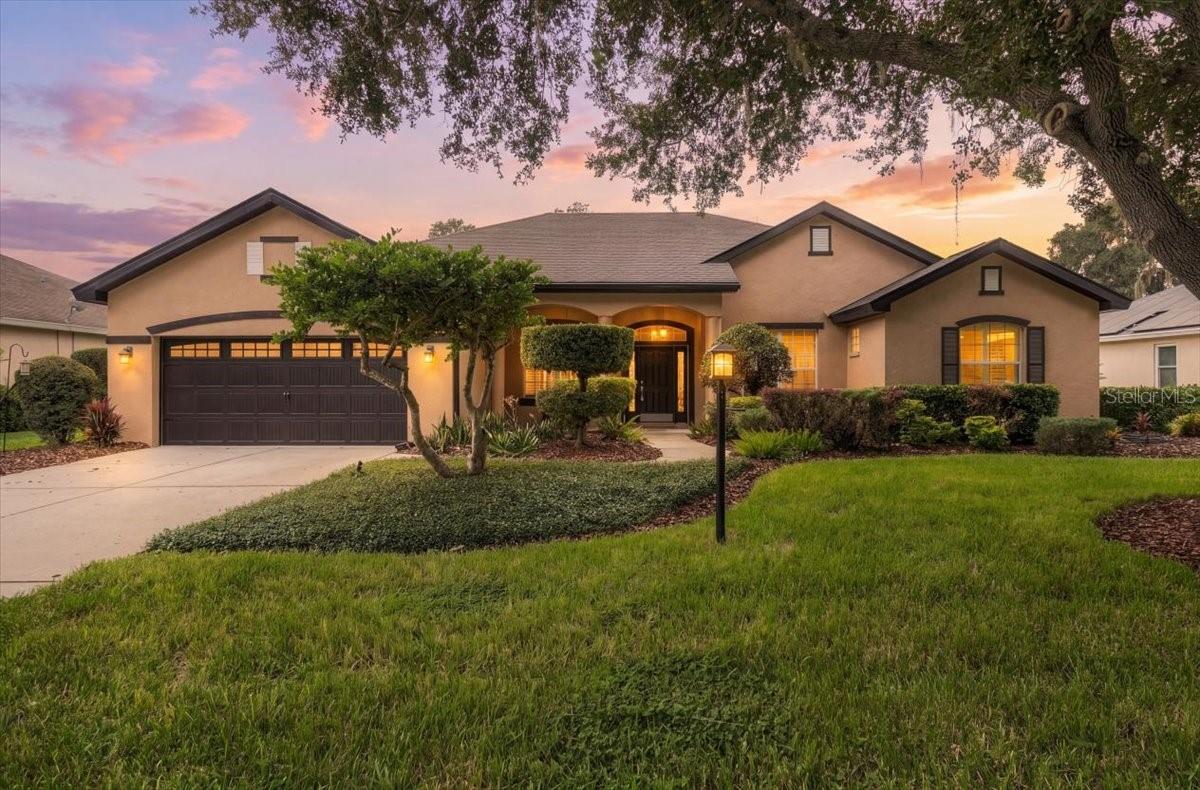 Tan stucco home with a double garage, arched entry, and landscaped lawn at dusk.