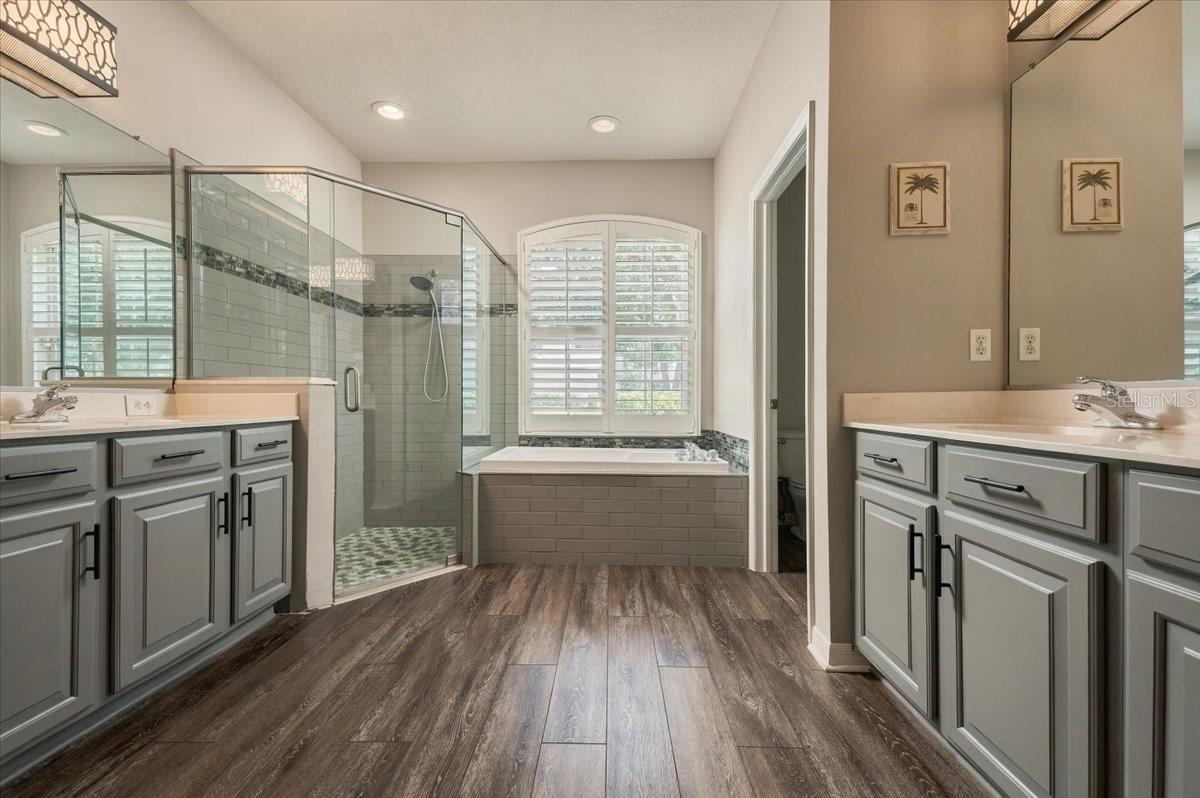 Bathroom with double gray vanities, glass shower, and soaking tub beneath shuttered windows.