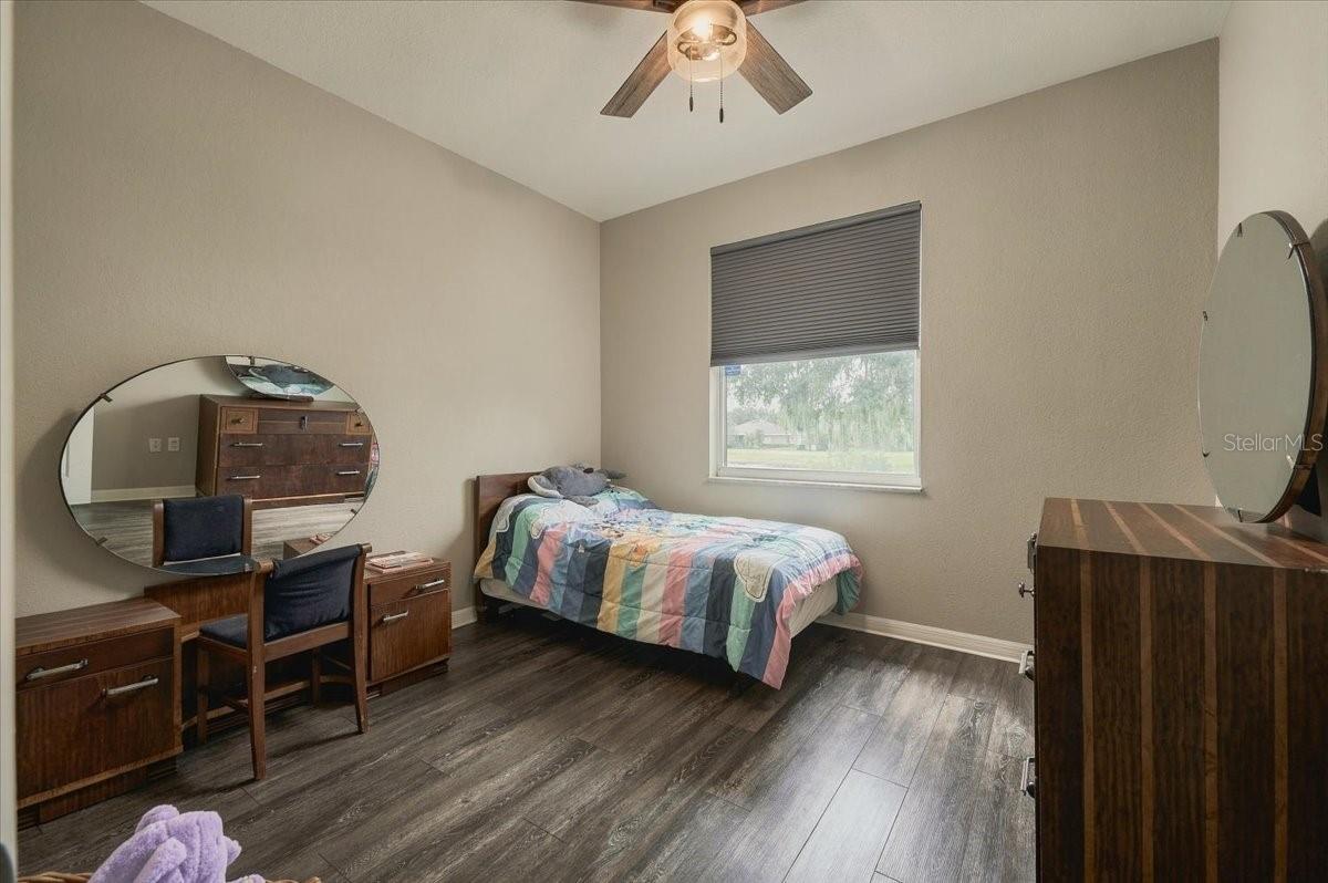 Furnished bedroom with a multicolored quilt, matching wood furniture, dark flooring, and a window with a lowered shade.