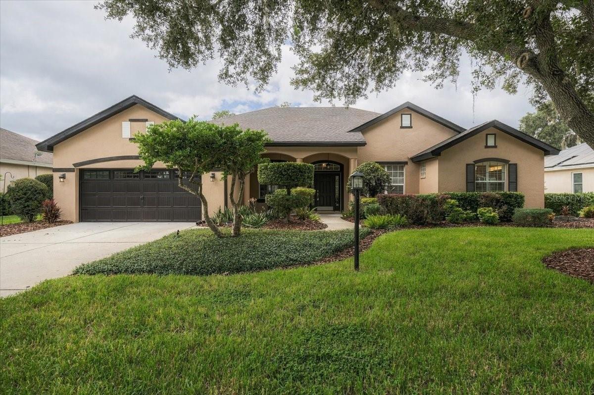 Tan stucco home with a double garage, covered entry, and landscaped front lawn beneath a large oak tree.