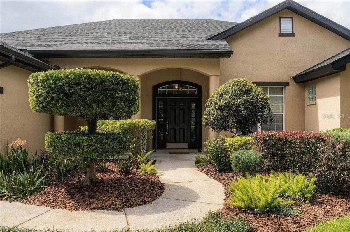 Home entrance with a black arched door, curved walkway, and manicured shrubs.