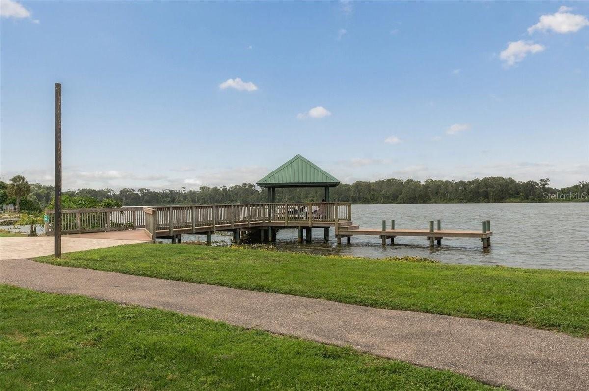 Wooden pier and green-roof gazebo extending over a lake beside a grassy lawn and paved path.