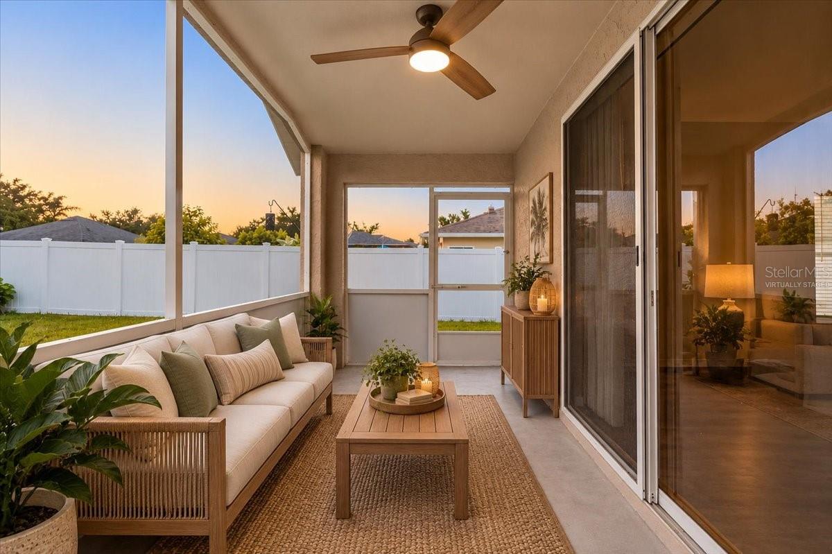 Screened patio with cushioned sofa, wood coffee table, ceiling fan, and white privacy fence at sunset.