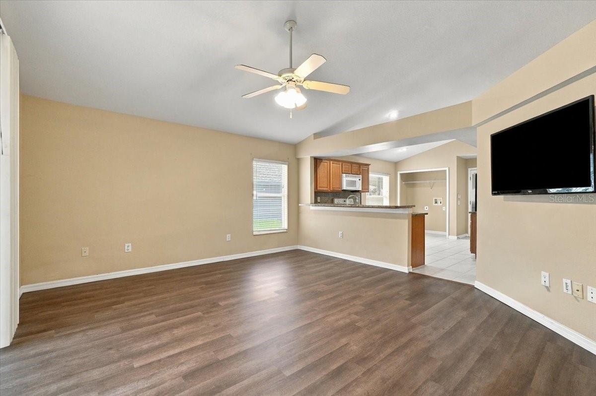 Living room with dark wood flooring, ceiling fan, wall-mounted TV, and open kitchen.