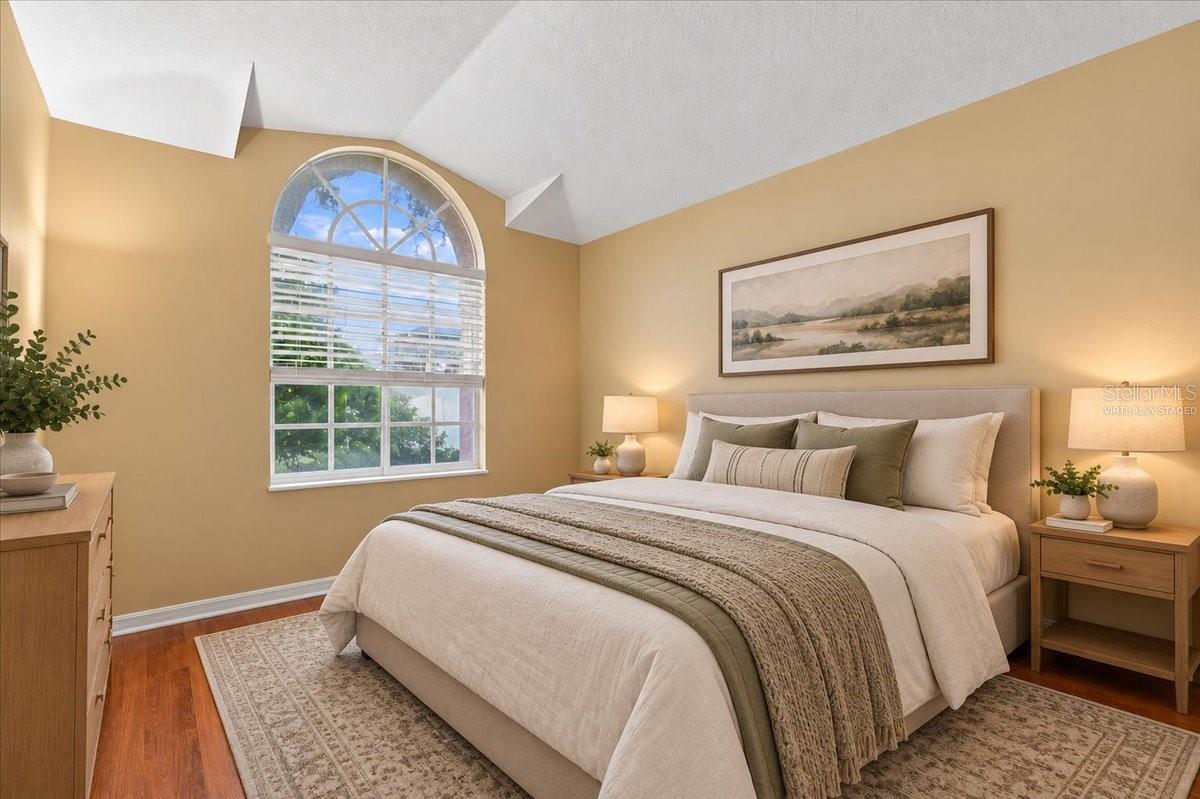 Staged bedroom with a king bed beneath a framed landscape, beside a tall arched window.