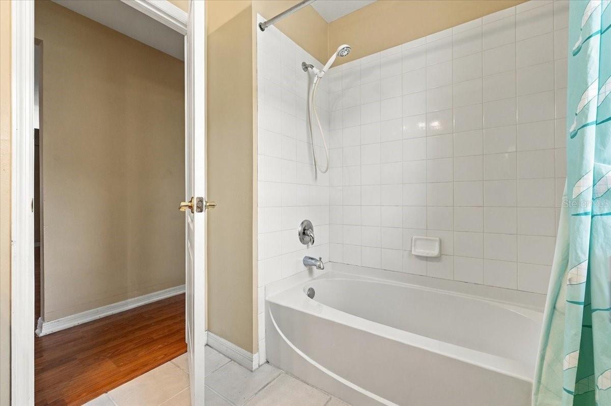 Bathroom with a white tiled tub and shower beside an open doorway to a wood-floored room.