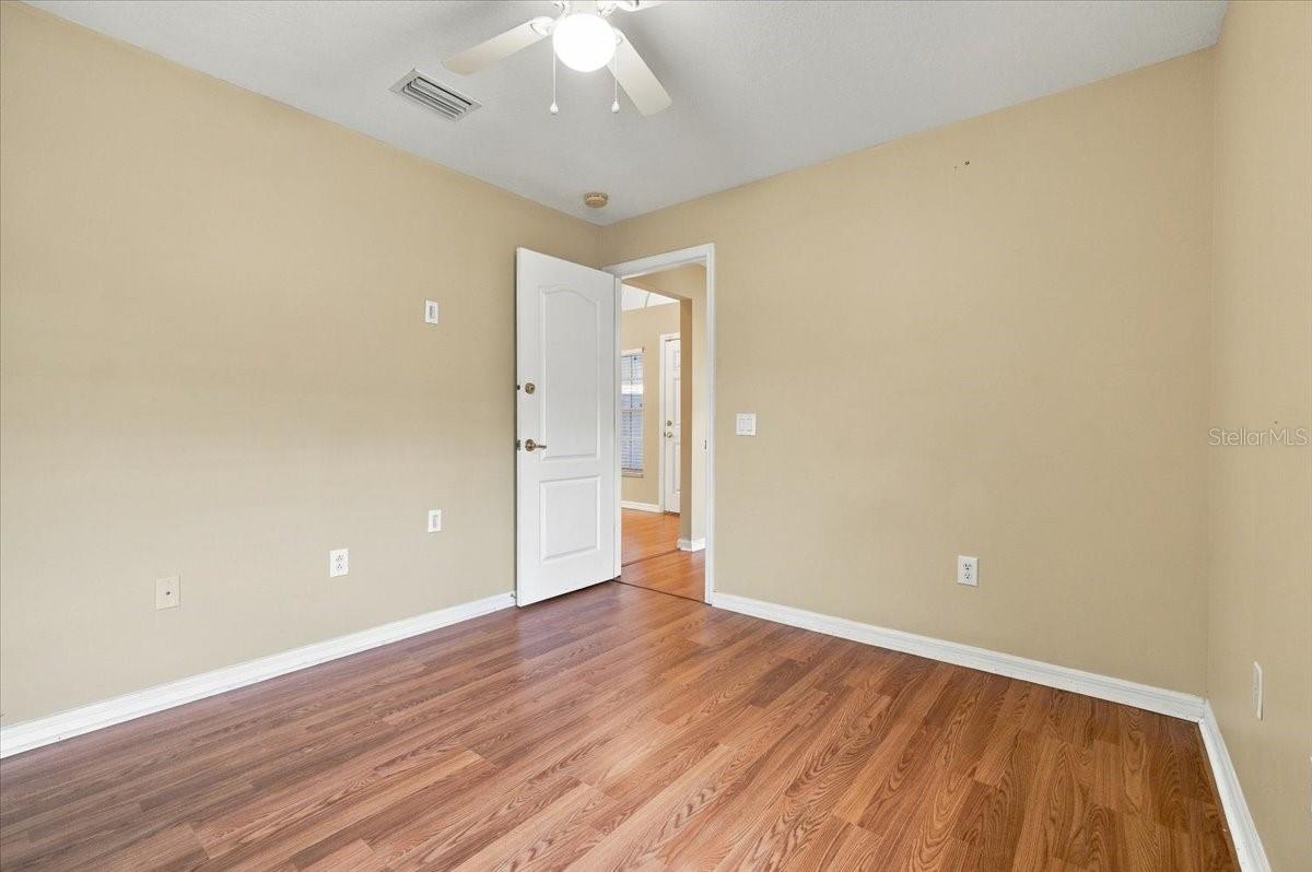 Empty room with wood-look flooring, beige walls, and a ceiling fan; an open door leads to another room.